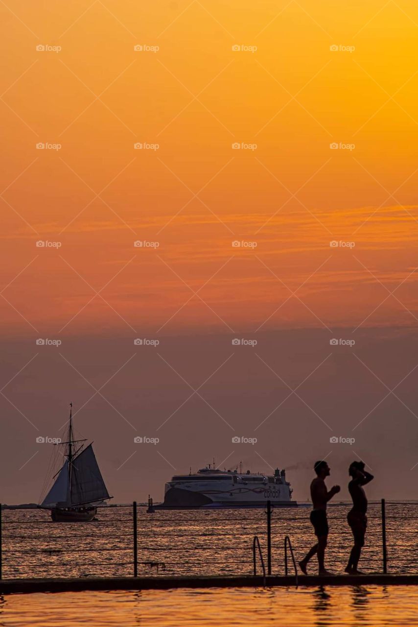 Young people on Saint-Malo mettalic diving platform with a saiboat and a cruise ship on the horizon, at sunset