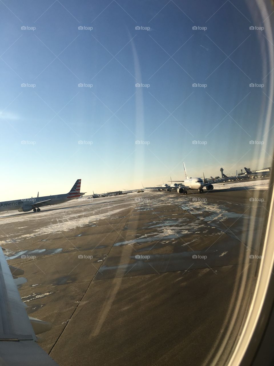 View from a plane window waiting to take off from the runway. There are a few planes on the runway waiting to take off. There are some ice from snow on the road.