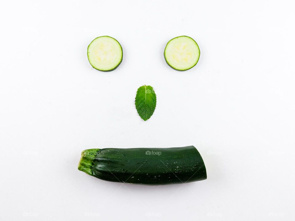 A green fresh zucchini is cut and folded in the form of a face with eyes, a nose, a mint leaf and a smile in the center on a white background, flat lay close-up. The concept of vegetables isolates, vegetables signs and symbols.