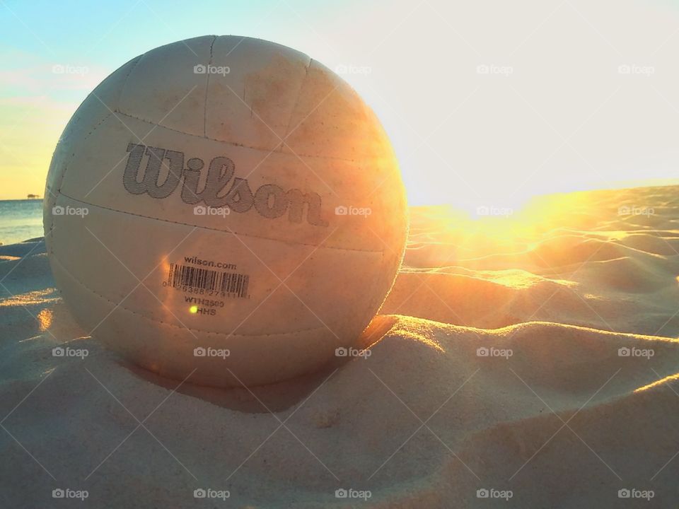 A Wilson volleyball laying gently in the sand on the seashore backlit by the bright setting sun