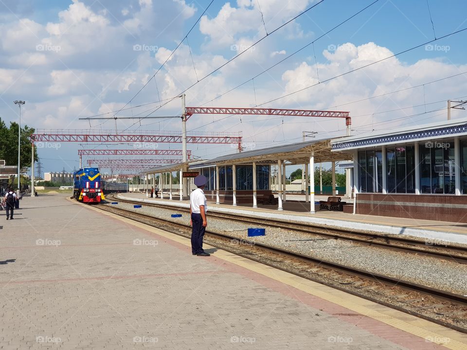 Bright colored train arriving at platform, where only one person, the station chief, is waiting.