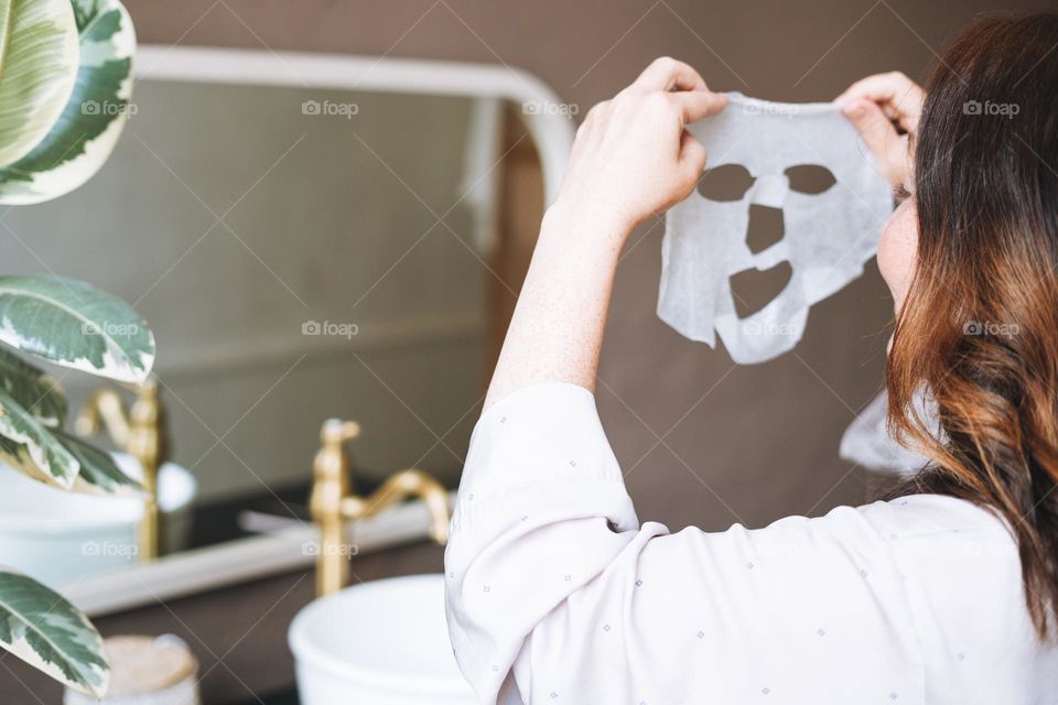 Adult brunette woman with sheet mask on her face in the bathroom at home