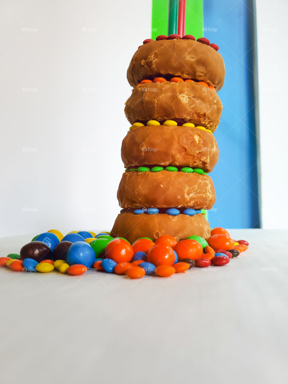 Five stacked donuts slightly leaning to the right & decorated with M&M minis. Surrounded by peanut M&Ms on a white surface with a blue, green and white solid background.  Reminiscent of the classic Fisher Price rock-a-stack infant toy.