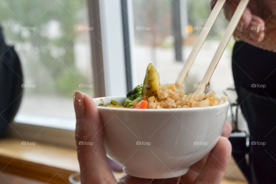 A closeup of a woman's hands using chopsticks to eat from a small bowl of traditional Chinese food comprising vegetable stir fry and rice