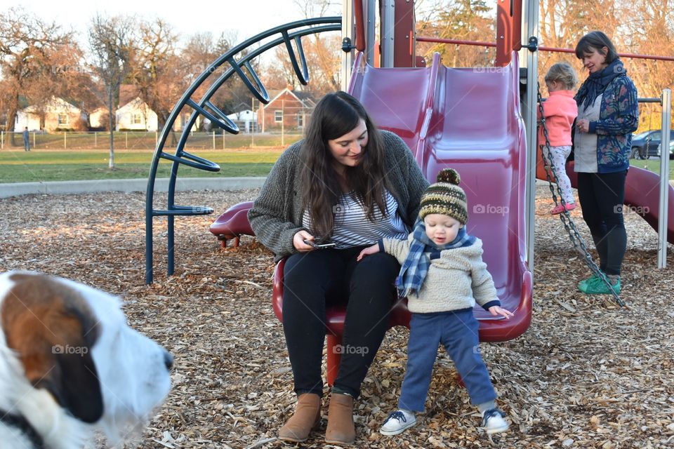 Cute toddler boy playing at playground with his mother while his dog watches 