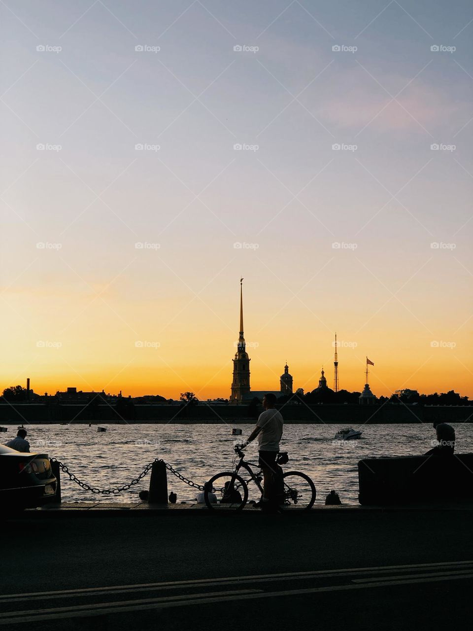 Young male with bicycle standing on pier near water, looking at beautiful sunset, Petersburg, Russia 