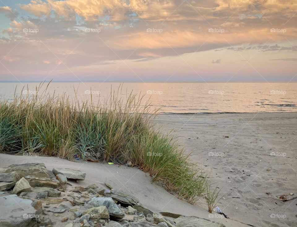 Liquid Fresh water Lake Eerie on Cedar Point shoreline with beach grass in the foreground surrounded by a sandy, rocky beach with purple pink and blue sunset sky divided by a white puffy cloud 