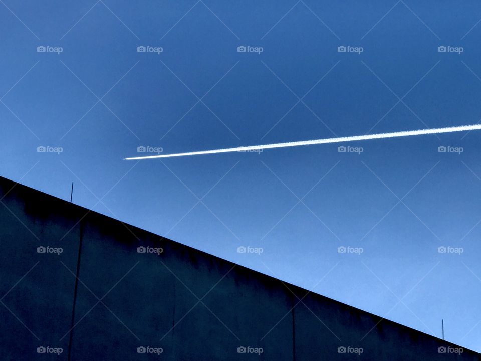 Airplane against a blue sky is leaving a white stripe of condensation trail,above a building