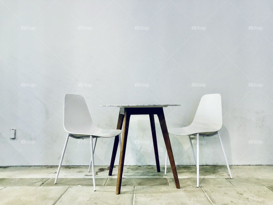 Simple cafe table and two white chairs against a white wall. Minimalist view emphasizing the color white.