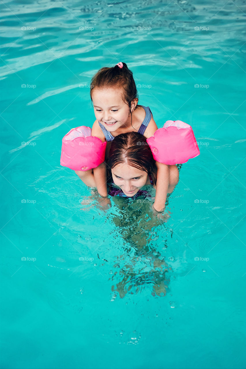 Young woman teaching to swim her younger sister and playing with her in swimming pool. Candid people, real moments, authentic situations