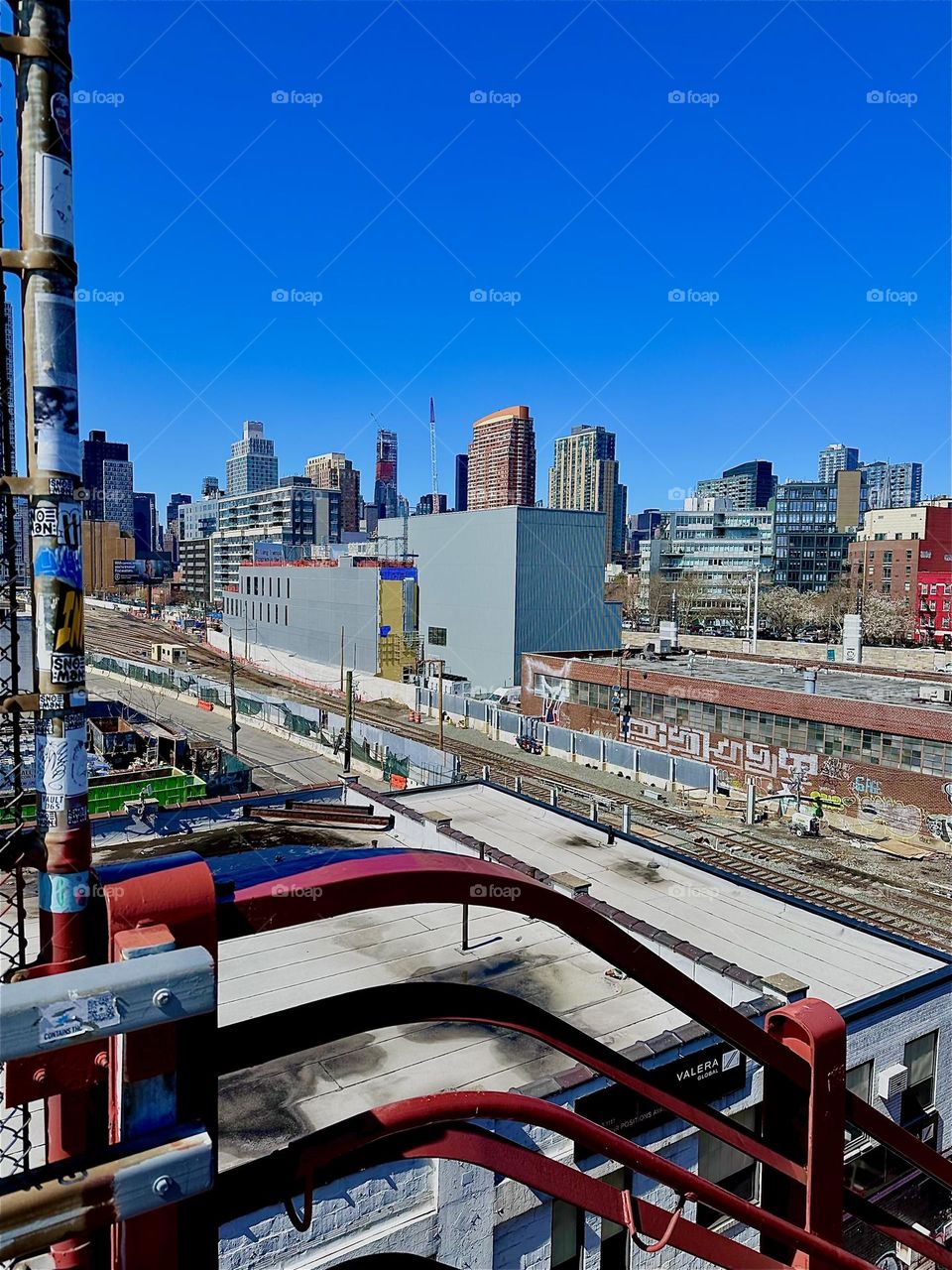 From the top of the red metal staircase of the “Pulaski Bridge” at “Newtown Creek” in LIC, Queens we see the train tracks of the “Long Island Railroad” and the building that houses “iFly”, an indoor flight simulator. 2024. Hypnotic Productions