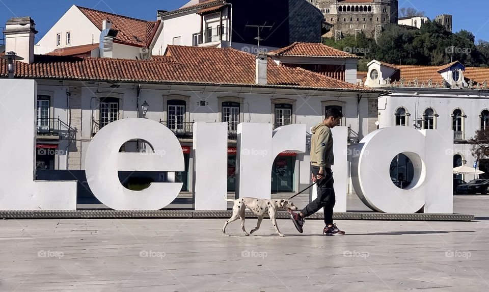 A man walks his Dalmatian in the city of, past a large white sign with the name of the city, Leiria.