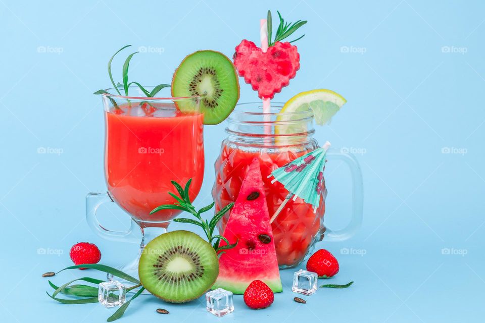 Multivitamin drink in glasses with fresh berries, fruit pieces, rosemary sprigs, ice cubes, straws and skewer paper umbrella on a blue background, close-up side view.