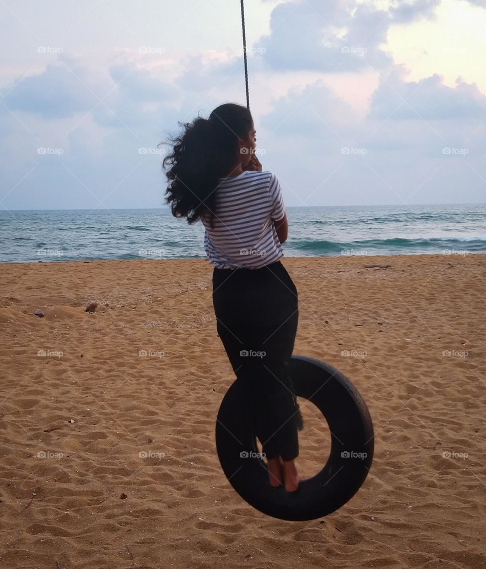 This is a capture of scene a sea beach.The girl is playing on a tire that is hang with a rope. There are crashing waves in the sea. The cloudy sky with the setting sun is the background of the capture. The sand is golden colour .