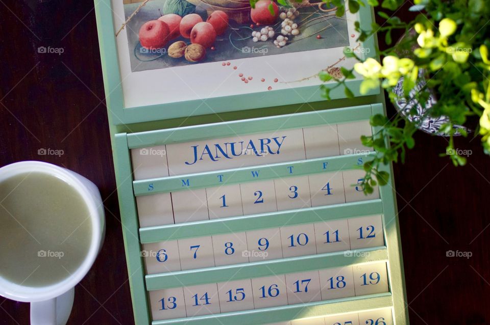 Green wooden perpetual calendar with interchangeable ivory-colored date tiles and light blue lettering, matcha latte in a white cup, and a small plant partially illuminated by sunlight on a dark wood surface 