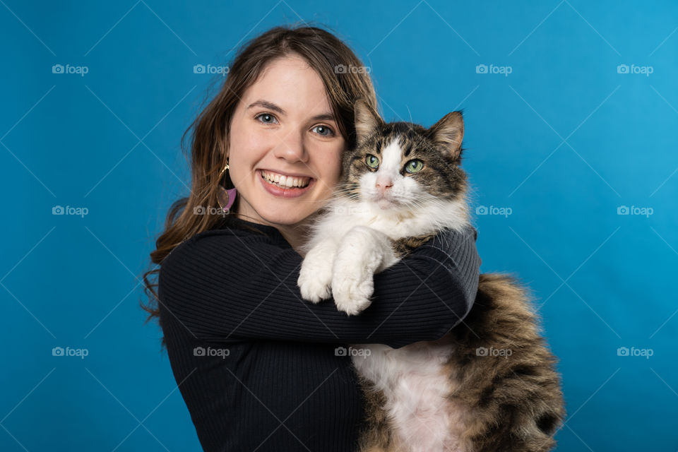 A young woman hugs her Maine coon cat happily in front of a blue backdrop 