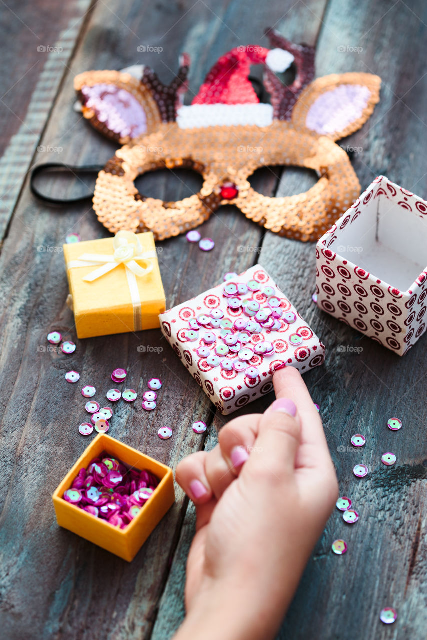 Close shot of female hand decorating Christmas reindeer mask with sequins following diy ideas on wooden table