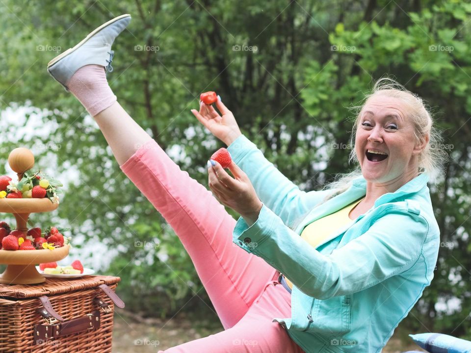 Beautiful caucasian young and happy blond girl having fun lifting up her leg and holding a strawberry in her hands at a picnic on a summer day. Concept happy people, picnic time.