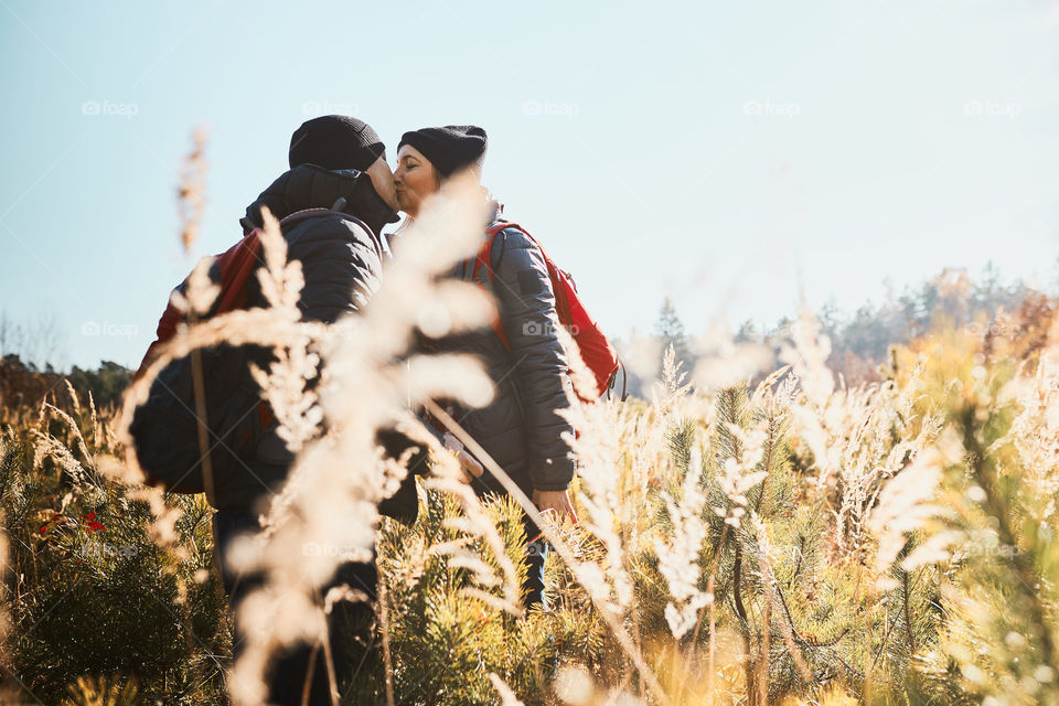 Couple sharing a passionate kiss while vacation trip. Hikers with backpacks standing on trail. People walking through tall grass along path in meadow on sunny day. Active leisure time close to nature
