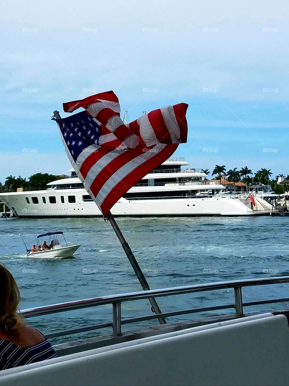 Boat flying American Flag in river with yacht & small boat in background. It's windy with blue sky!