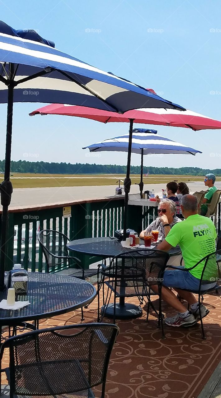 Umbrella covered outside restaurant, airport for small planes, blue sky, runway for watching.