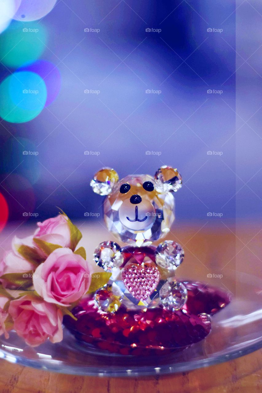 Teddy bear with a pink heart. Glass statuette in a romantic setting. Multicolored bokeh on blurred background.