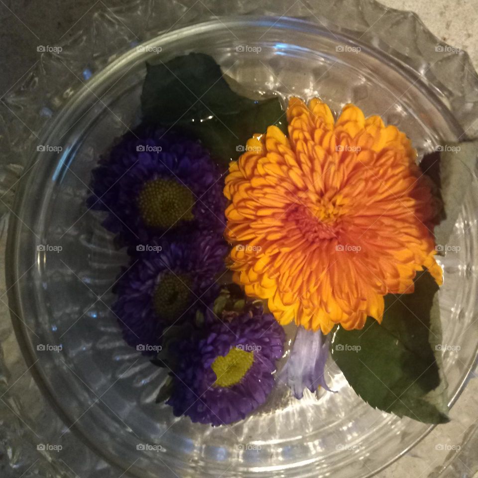 Large orange and white mum with green foliage and lilac purple flowers in an ornate crystal dish as a centerpiece.