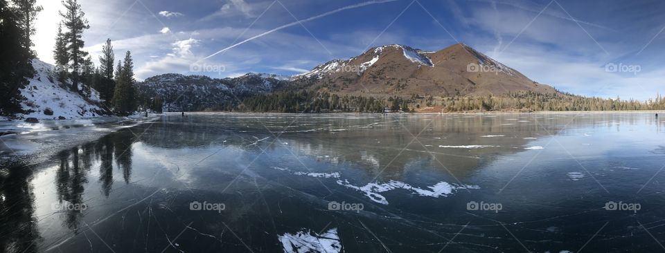 A panoramic view of mountains roughly reflected in the ice of a frozen over lake in the sierras. 