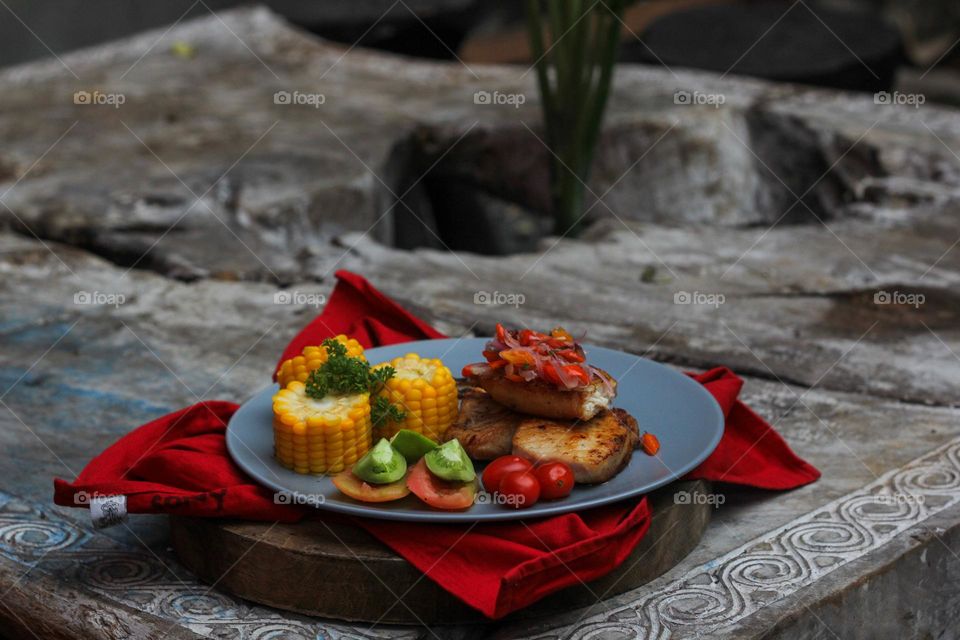 Tuna steak cooked with corn garnished with cherries, sliced tomatoes and lemon topped with sliced hot chili peppers on a baby blue melamine plate lined with red cloth and thick wood. Restaurant food.