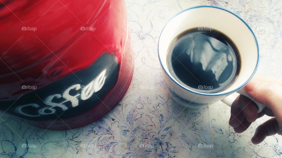 Coffee canister or red crock with coffee written on it in chalk and a warm cup of Joe being held by a woman's hand