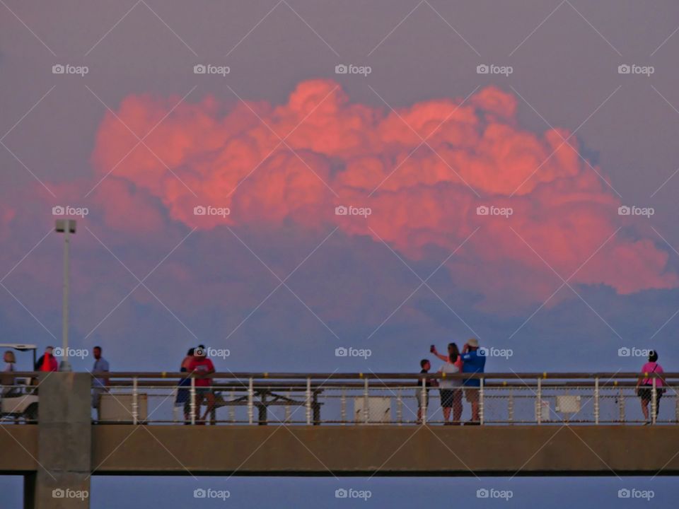 Pink clouds radiating over the fishing pier from the rays of a dramatic sunset. People are taking selfie’s while enjoying their vacation