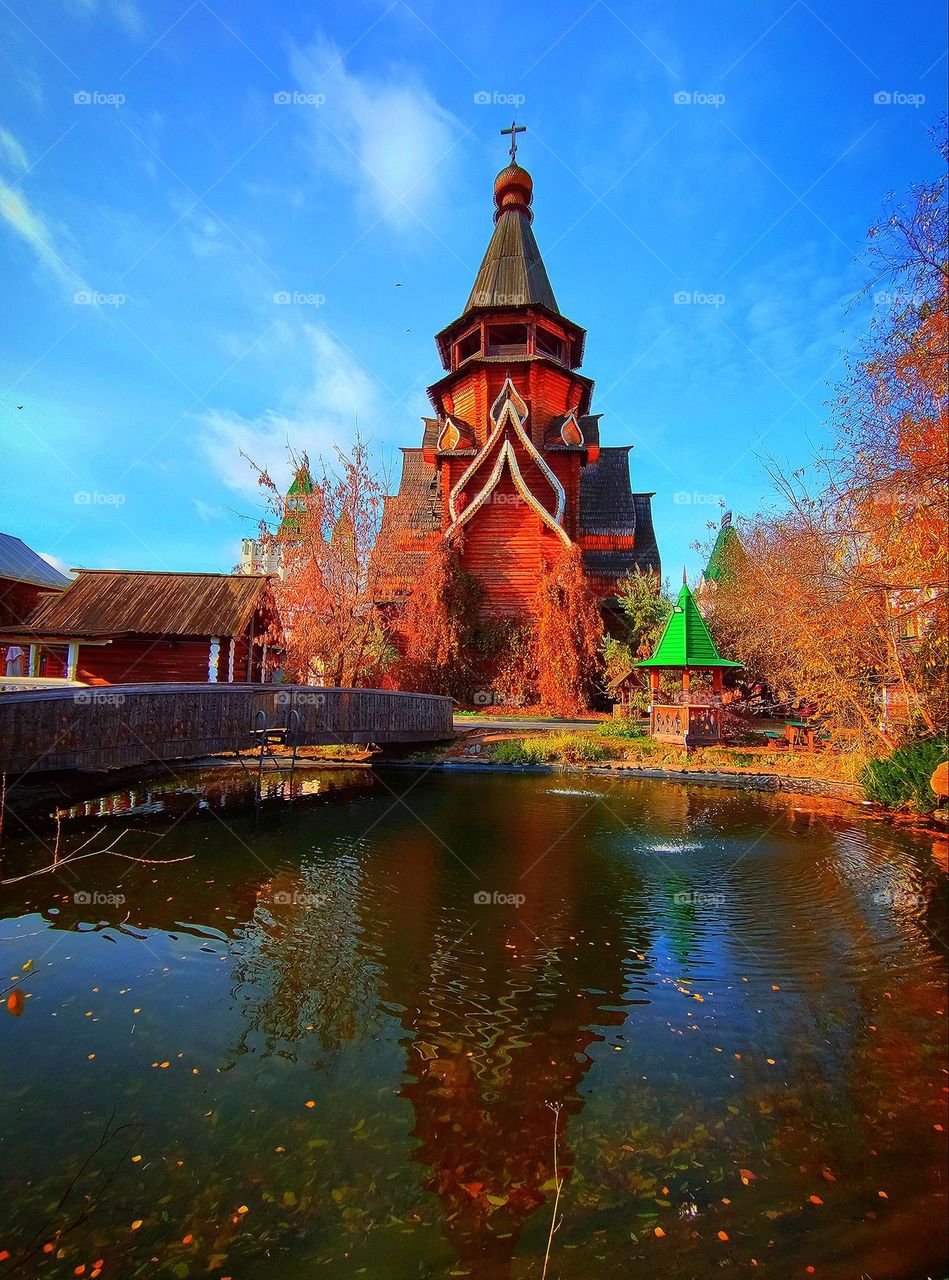 Wooden church, wooden bridge across the pond. There is a wooden well near the church. Near the pond there is a wooden gazebo. Around the autumn trees. Above the church there is a blue sky with white clouds. The church is reflected in the water