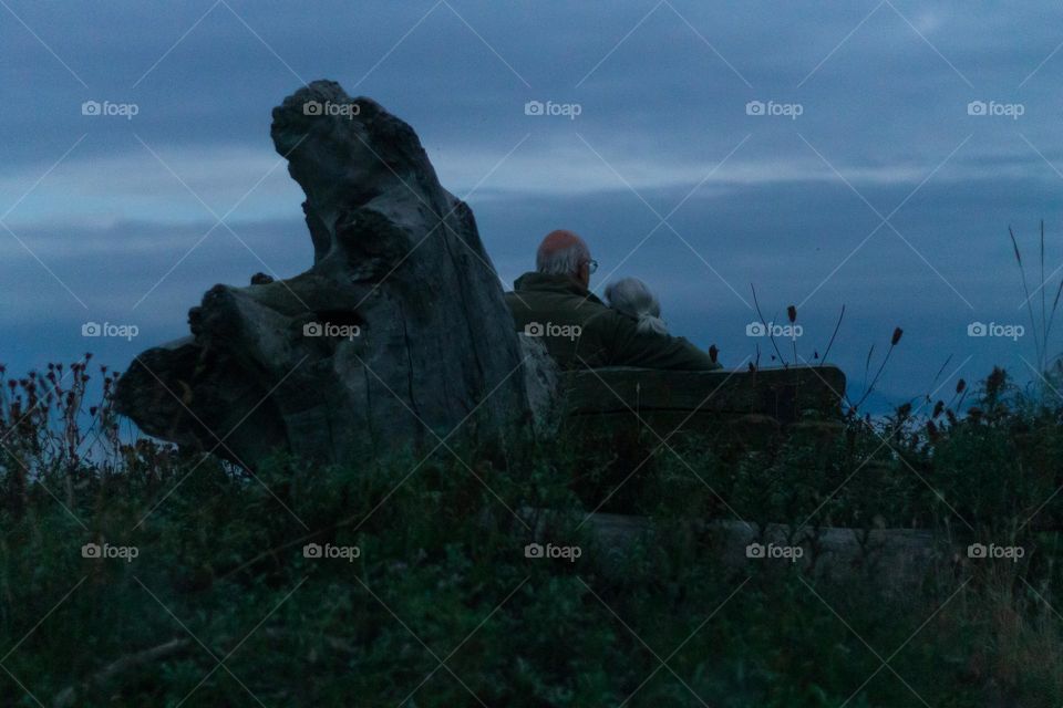 Darkening twilight shot of backs of a couple cuddling on a bench at the beach. Soaking in as much light as possible with super wide aperture and super long shutter speed and a tripod. Magic mesmerizing moment.