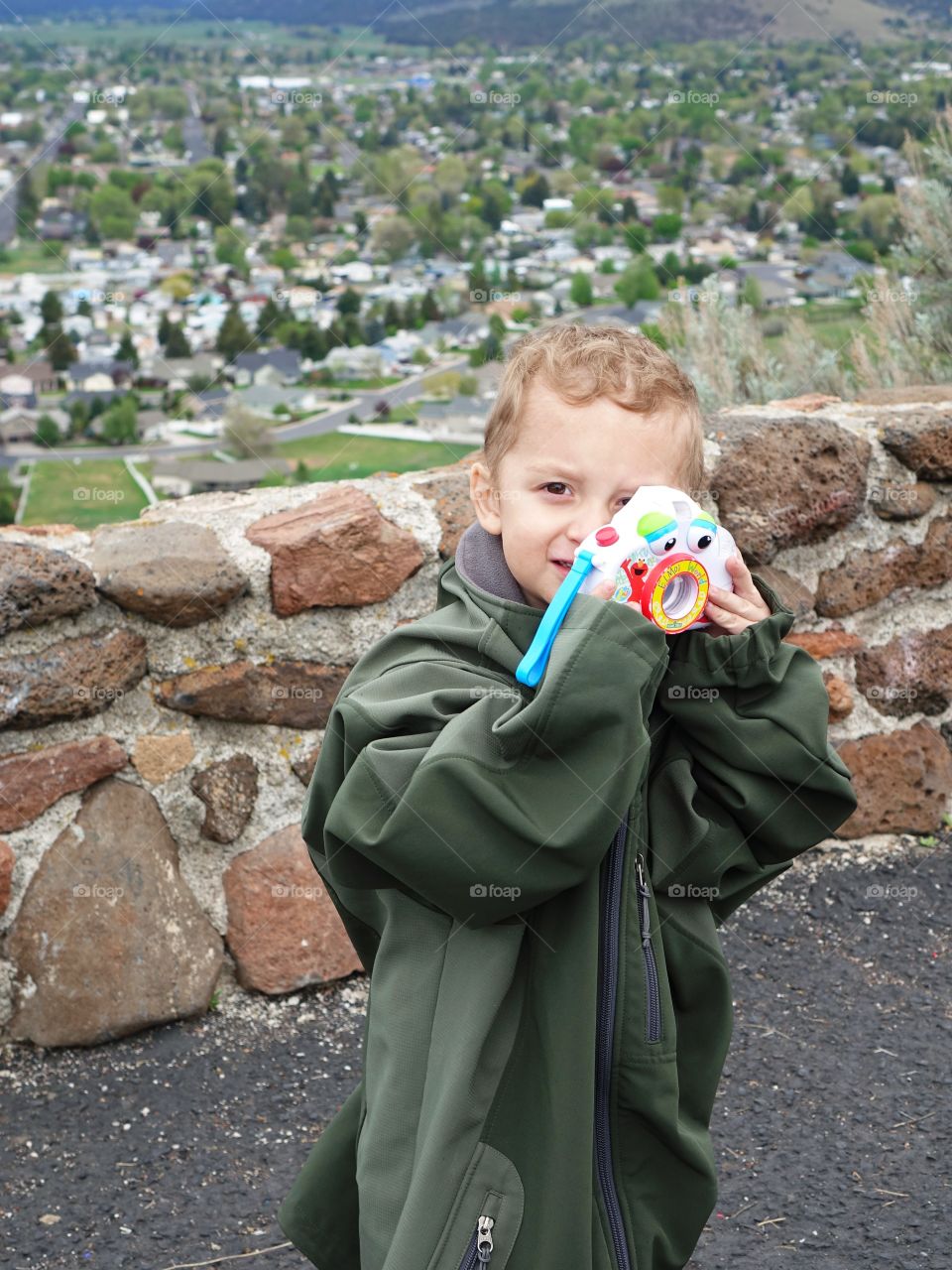 A cute young boy in an adult jacket that hangs to the ground takes a photo with his toy camera. 