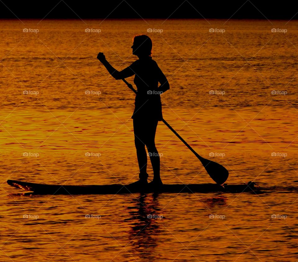 Silhouettes and shadows - A women does her daily exercise by paddle boarding in the bay as the sunset descends 