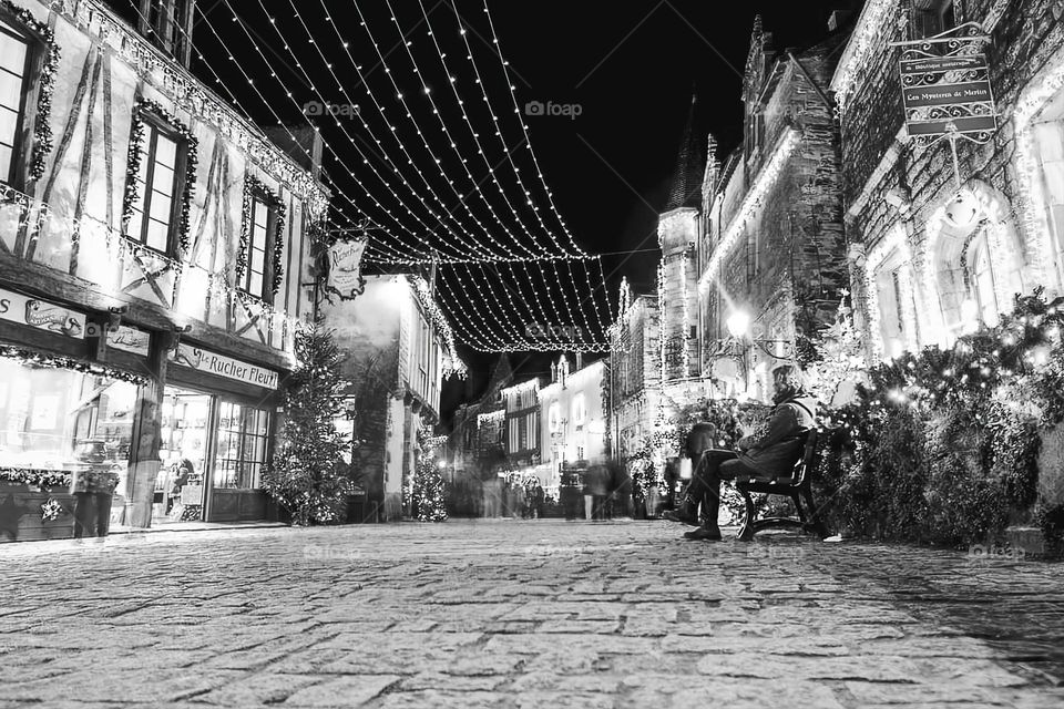 Black and white shot of a woman sat on a bench in the main street of Rochefort en Terre adorned with its christmas decorations and entirely iluminated by the lights behind the shops and houses' windows, at night time