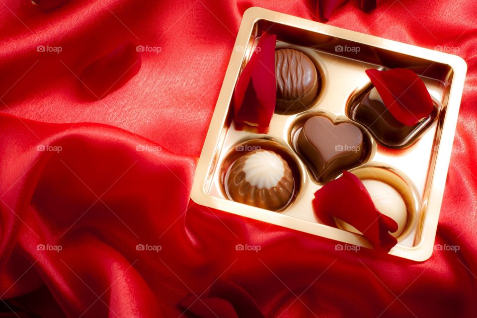 Golden box of differently shaped chocolates on red silk background surrounded by rose petals for Valentines's day 