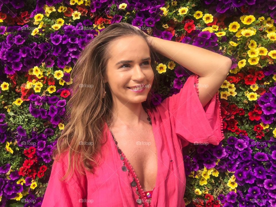 Portrait of pretty young woman wearing pink dress surrounded by wall of multicoloured flowers