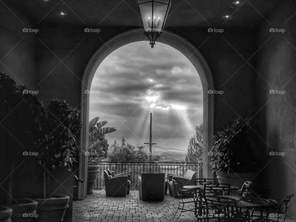 Peering out from a balcony with the sun shining through the clouds with chairs placed in view and trees in the outer frame. A cross in center frame. In black and white.