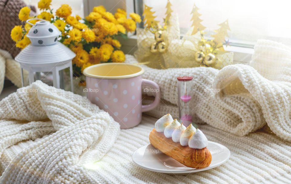 Still life made by more objects like mug, flowers, eclair, fabric make cozy atmosphere.