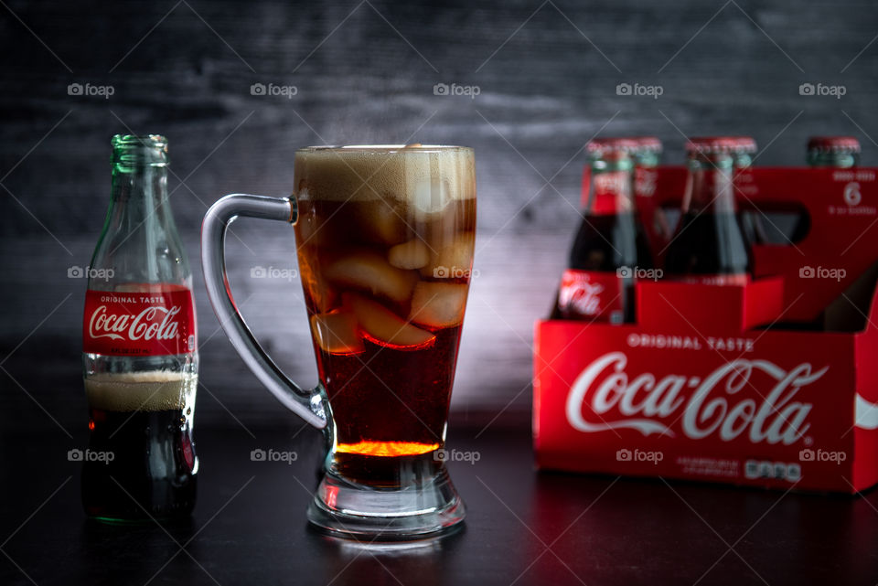 Backlit tall glass of Coca-Cola over ice next to a six-pack