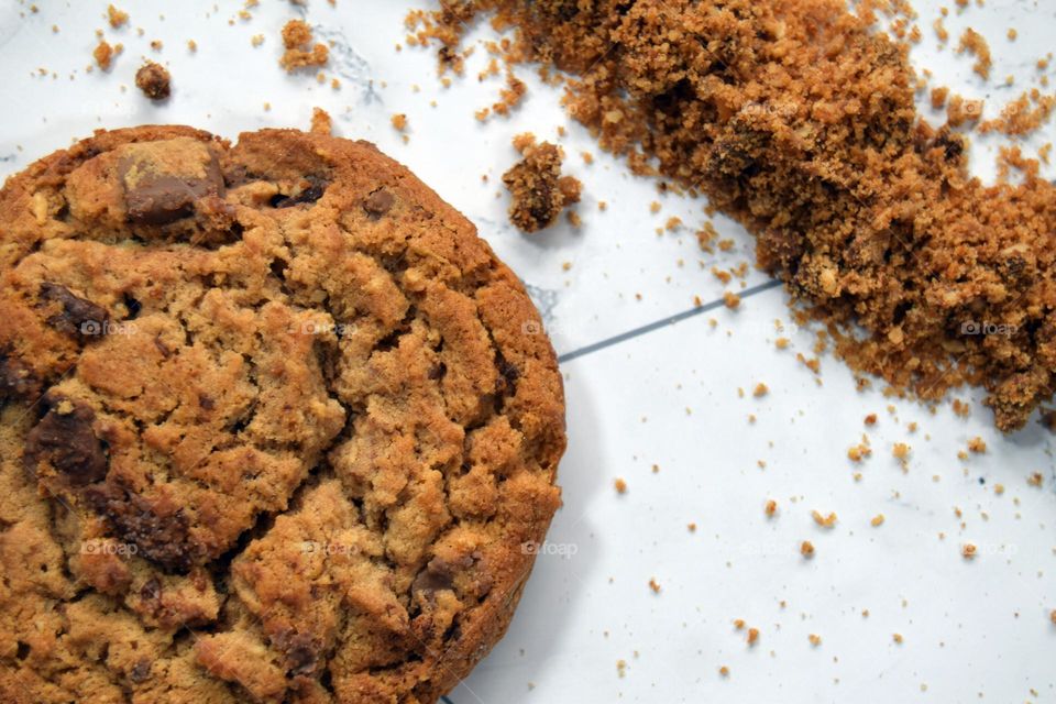 A chocolate chip cookie on a white marble flatlay background accompanied by a sprinkling of chocolate chip cookie crumbs.