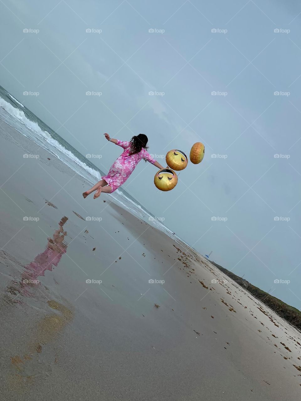 Little Girl Seems To Be Flying And Carried Up In The Air By The Three Helium Emoji’s Balloons On The Beach By The Ocean.