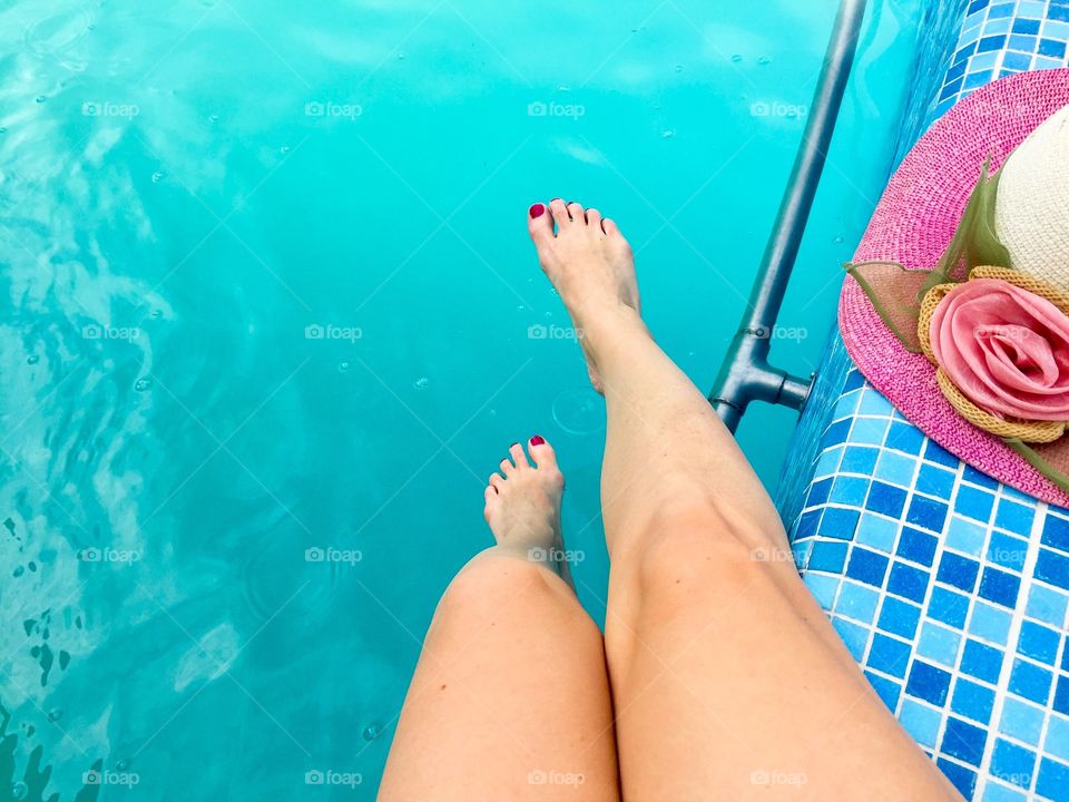 Woman's legs in the swimming pool with pink summer hat beside