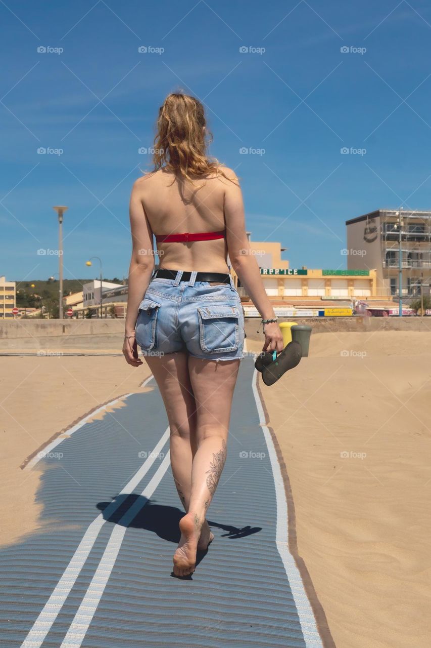 Young blonde woman walking on the beach wearing jeans shorts and holding flipflops in her hands