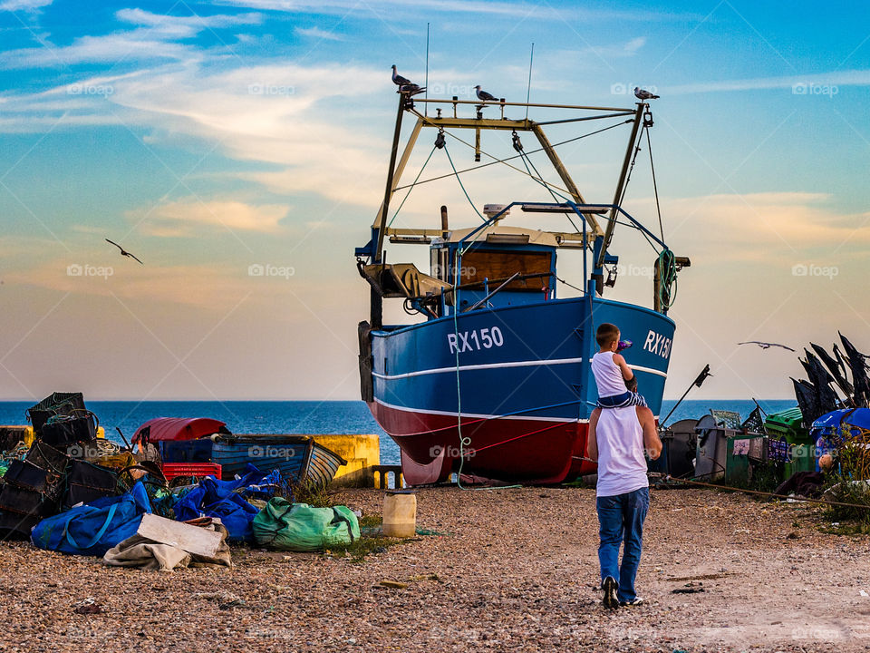 A man walks with a child on his shoulders past the working fishing beach at Hastings 