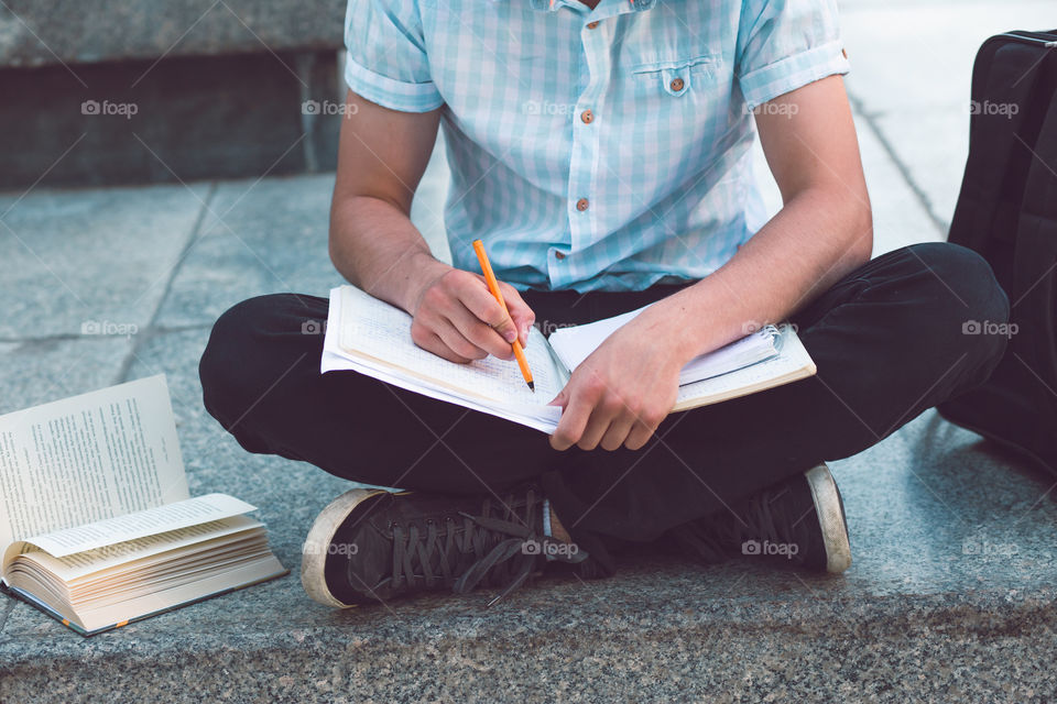 Student making the notes learning from books sitting on a monument outside of university. Young boy wearing a blue shirt and dark jeans