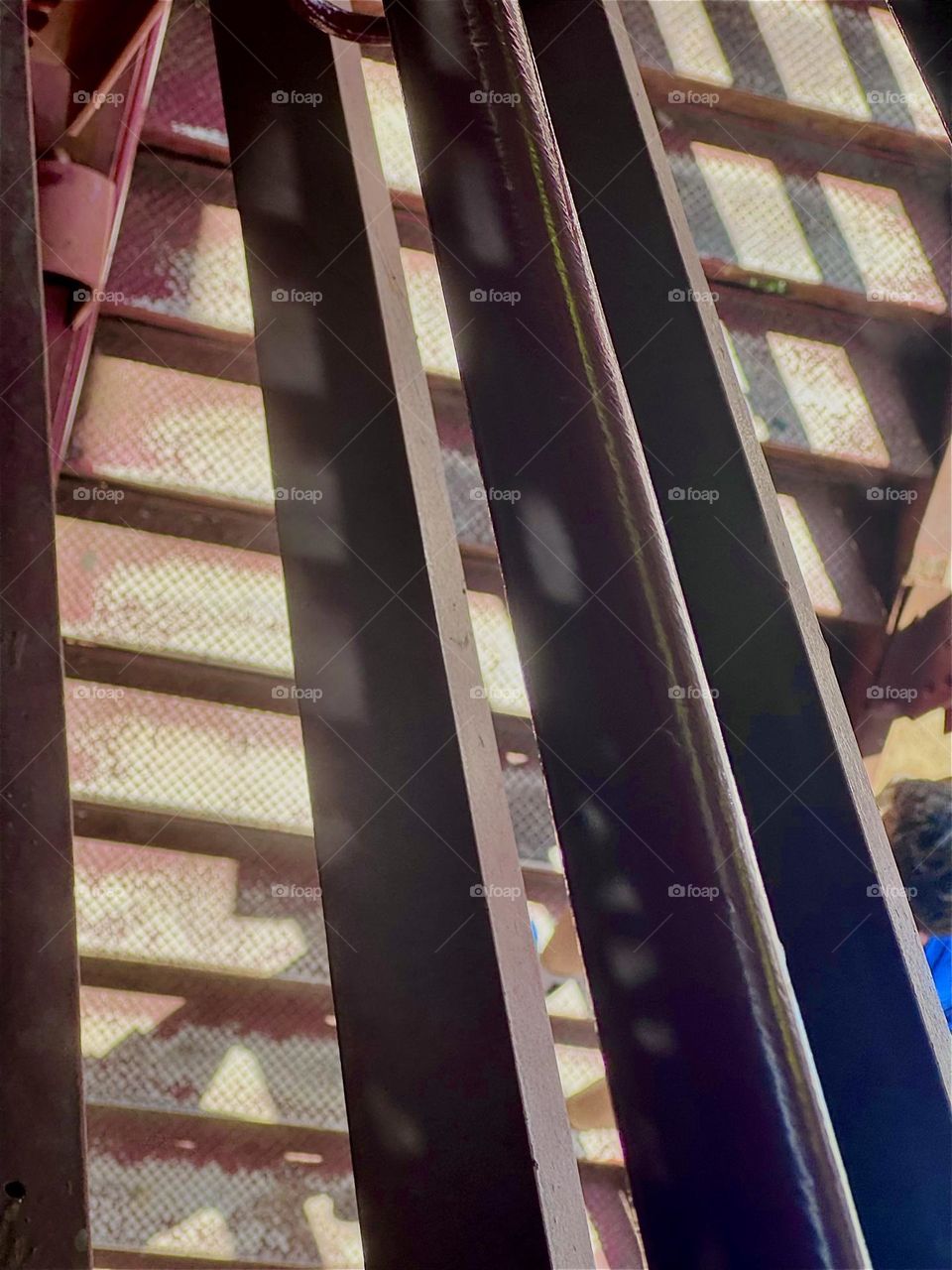 These are the steps of the red metal staircase of the “Pulaski Bridge” at “Newtown Creek” in LIC, Queens seen through the railing from above on a bright sunny afternoon in late May of 2024. Hypnotic Productions 