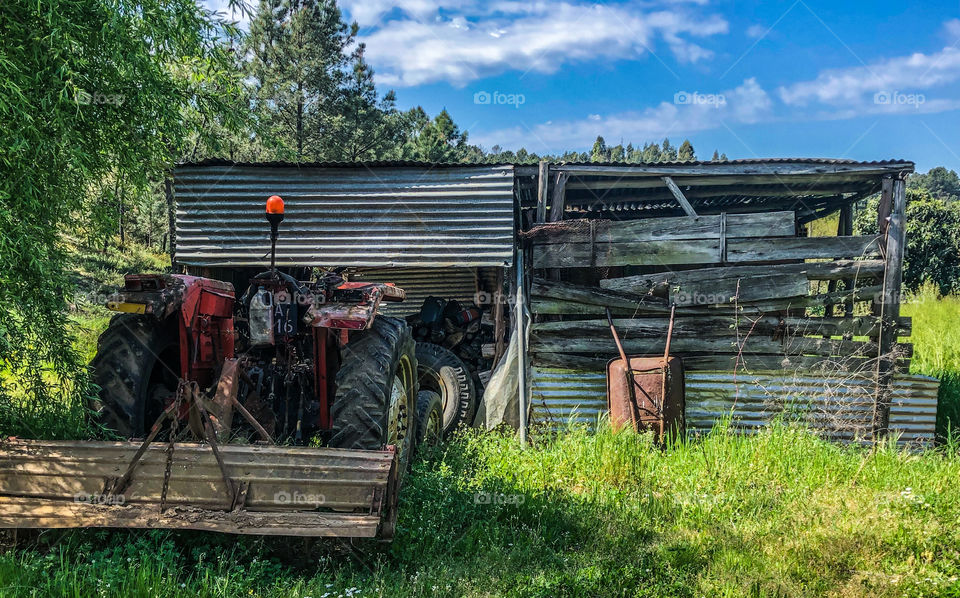 A shack constructed of mostly corrugated metal sheets houses a tractor and a rusty wheelbarrow leans up against the shack in this countryside scene