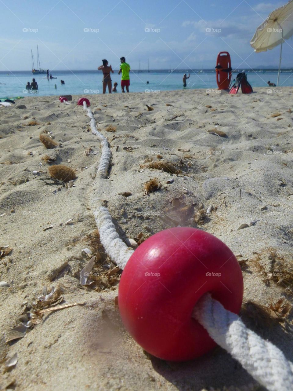 Panoramic view of a sandy beach seen at ground level. In the foreground, a small red buoy is attached to a rope, arranged towards the sea. A lifeguard is talking with a swimmer, an umbrella and lifeguard equipment on the right. Majorca
